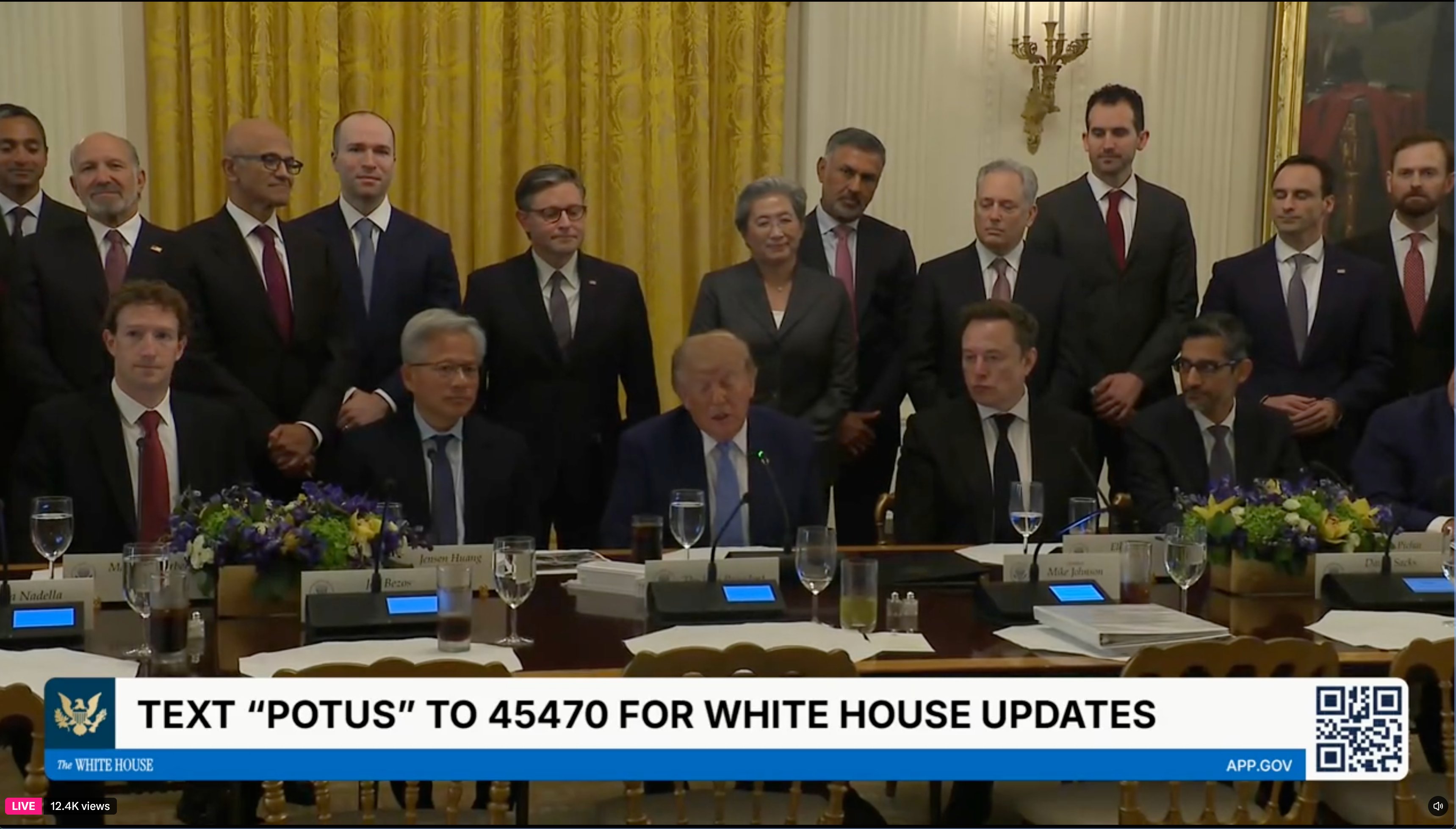 A group of men and one woman stand behind a conference table where Donald Trump is seated at a microphone. The table holds nameplates, glasses, papers, and flower arrangements. A White House banner reads “TEXT ‘POTUS’ TO 45470 FOR WHITE HOUSE UPDATES,” with “The WHITE HOUSE” and “APP.GOV”; a QR code is at the right. A “LIVE” badge and “12.4K views” appear at the lower left.